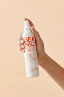 Hand holding a bottle of 'Sea Salt Texture Spray' by Eleven Australia against a beige background - hair Care Canada